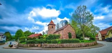 Zu sehen ist eine Sandsteinkirche mit Kirchtum auf einer leichten Anhöhe. Vor der Kirche wachsen Büsche und rechts neben der Kirche steht ein Baum.