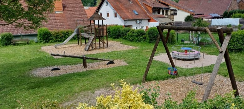 Zu sehen ist ein Spielplatz mit einer Schaukel, einem Kaurusell, einer Wippe und einem Kletterturm mit einer Rutsche. Im Hintergrund befinden sich Häuser.